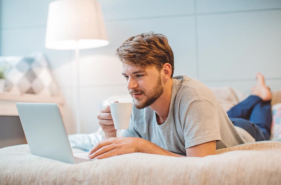 Mies makaamassa sängyllä juomassa kuumaa juomaa ja samalla lukemassa sähköposteja kannettavalta